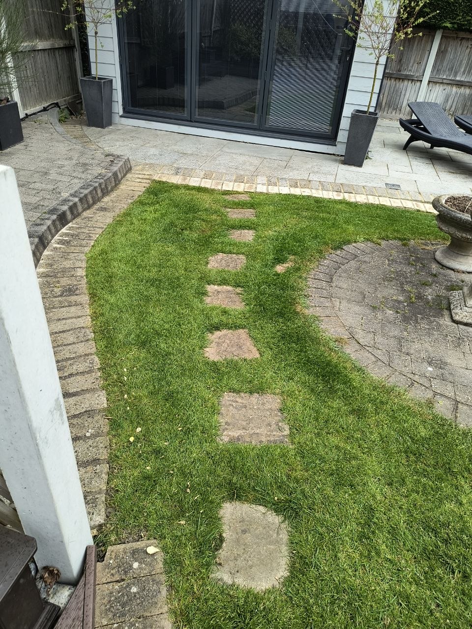 A side passage in Ash with a narrow lawn panel running between block paving and stepping stones leading from sliding glass doors, the grass sitting flush with the paving on a clean edge.